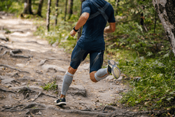a person in mid-stride, running on a rocky trail in a natural setting. They’re wearing grey compression socks, dark shorts, and a t-shirt suitable for physical activity. The compression socks may be for athletic performance or medical reasons.