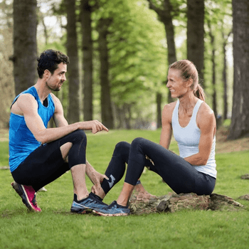Two individuals in athletic wear sit on the grass in a park, engaged in conversation, possibly taking a break from exercising.